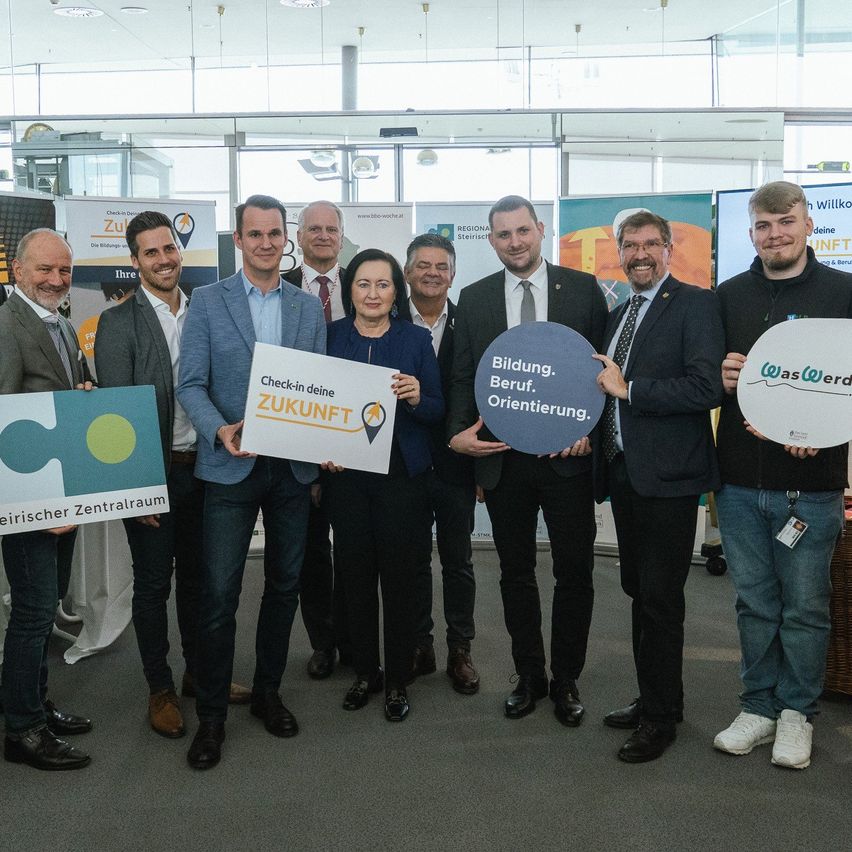 Eine Gruppe von Menschen posiert für ein Foto in einem Saal mit Schildern und hält Plakate mit der Aufschrift 'Check-in deine Zukunft' und 'Bildung. Beruf. Orientierung.' Sie lächeln und schauen in die Kamera.