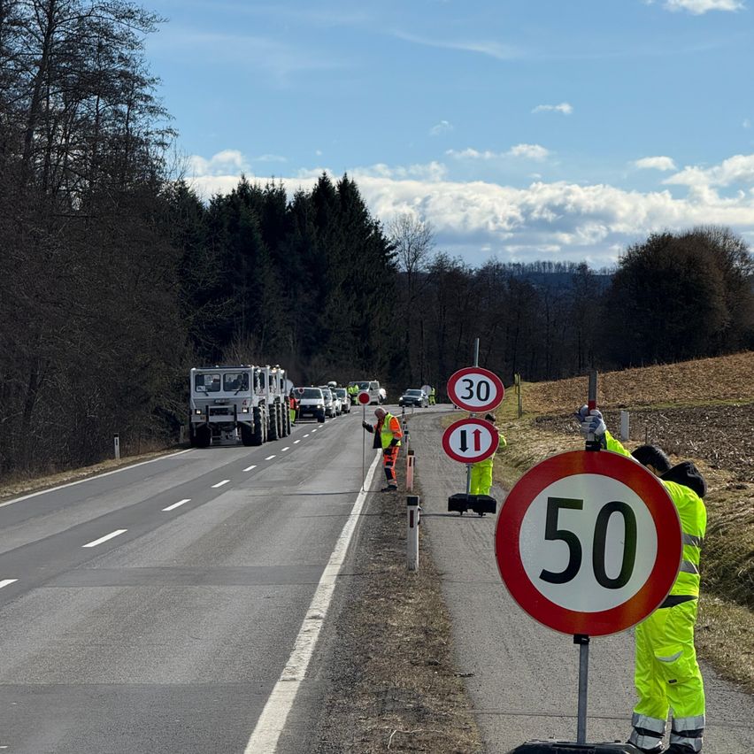 Eine Straßenwartungsmannschaft ändert die Geschwindigkeitsbegrenzungsschilder von 30 auf 50 km/h. Arbeiter in Warnkleidung sind in der Nähe der Schilder zu sehen. Die Straße wird von Bäumen und einem Grasfeld flankiert.