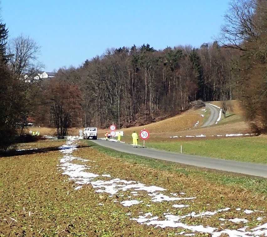 Eine Straße mit zwei Geschwindigkeitsbegrenzungsschildern. Arbeiter in Warnwesten stehen bei einem weißen Fahrzeug. Die Straße ist von Bäumen und Schnee umgeben. Ein Hügel ist im Hintergrund.