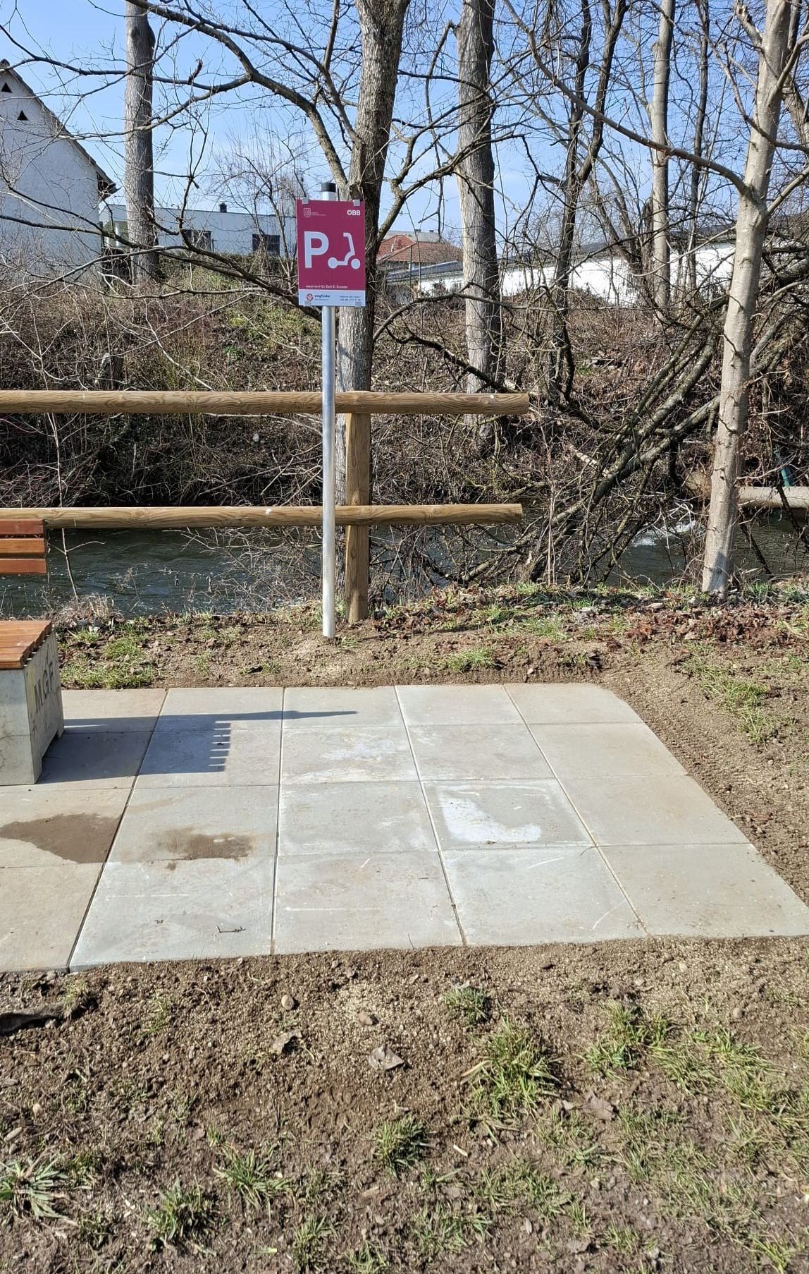 Eine Parkbank am Fluss, mit einem Schild, das auf einen Parkplatz für behinderte Fahrzeuge hinweist, umgeben von einem Holzzaun.