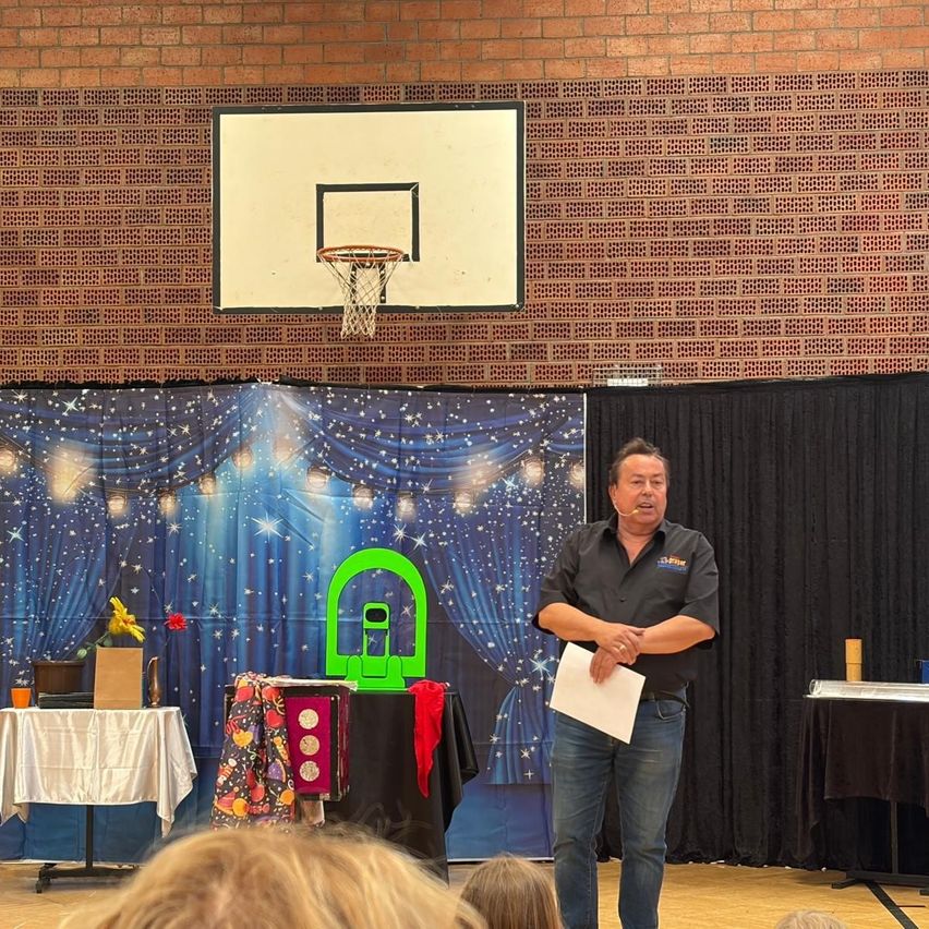 Ein Mann steht auf der Bühne mit einem Mikrofon, hält ein Papier in der Hand und eine Kulisse mit Sternen und einem Basketballkorb hinter ihm.