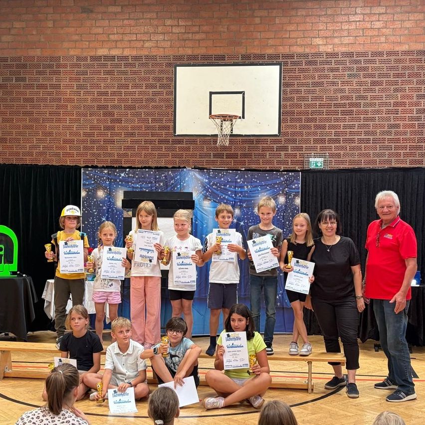 Eine Gruppe von Kindern und Erwachsenen steht auf einer Bühne in einer Turnhalle, hält Urkunden und Trophäen. Sie sind umgeben von einer Ziegelwand, einem Basketballkorb und einer Kulisse.