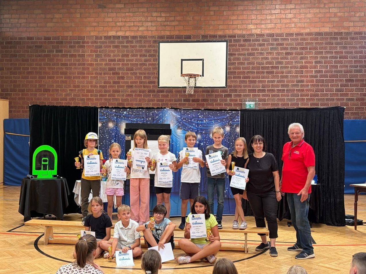 Eine Gruppe von Kindern und Erwachsenen steht auf einer Bühne in einer Turnhalle, hält Urkunden und Trophäen. Sie sind umgeben von einer Ziegelwand, einem Basketballkorb und einer Kulisse.