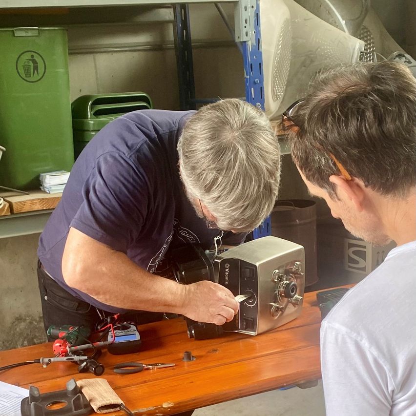 Zwei Männer arbeiten in einer Werkstatt an einer Maschine. Ein Mann hält ein Werkzeug und der andere beobachtet.