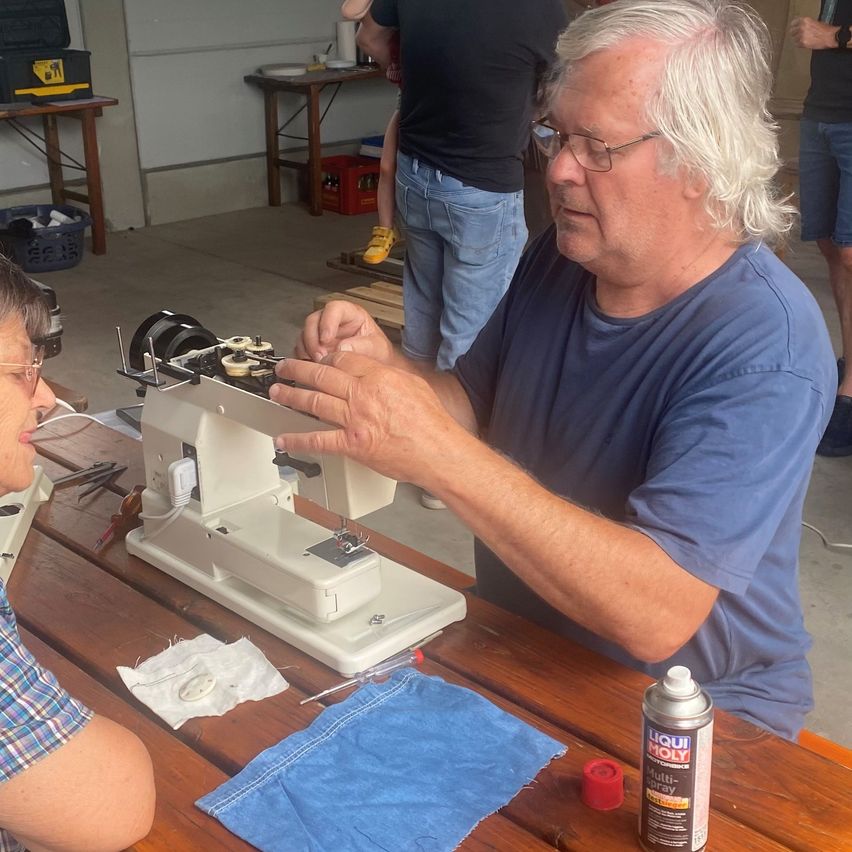 Ein älterer Mann mit Brille repariert eine Nähmaschine auf einem Holztisch, während eine Frau in einem karierten Hemd zuschaut.