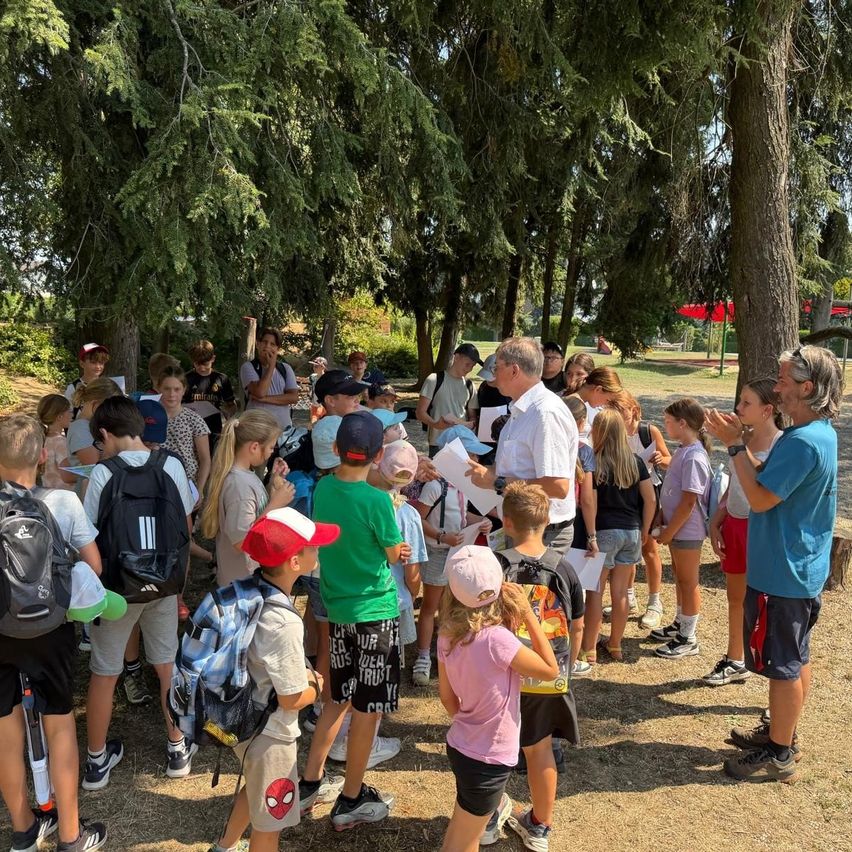 A group of children and adults gather under trees, with some wearing backpacks and hats. A man in a white shirt stands in front, holding a piece of paper.