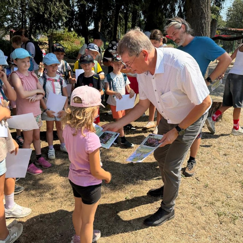 Ein Mann mit Brille und weißem Hemd gibt einer kleinen Mädchen in einer rosa Mütze Papiere in einem Park mit anderen Kindern und Erwachsenen.