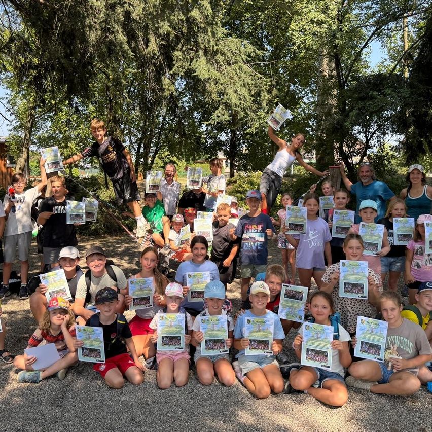 Eine Gruppe von Kindern und Erwachsenen posiert für ein Foto in einem Park, hält Urkunden und lächelt. Bäume und ein Gebäude sind im Hintergrund zu sehen.
