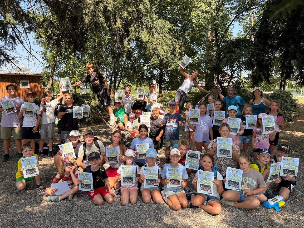 A group of children and adults pose for a photo in a park, holding certificates and smiling. Trees and a building are in the background.