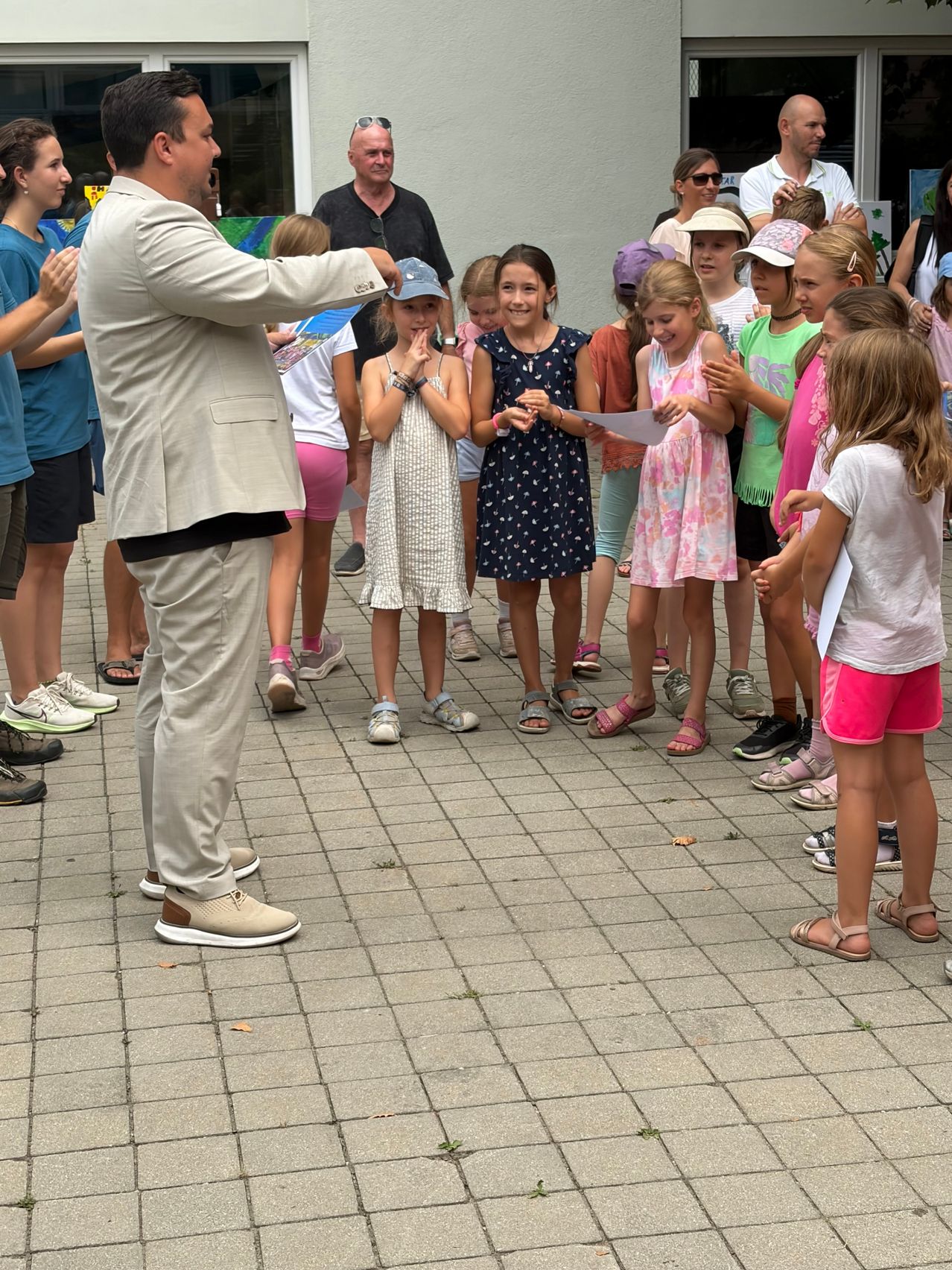 A group of children and adults stand in a circle on a brick floor, with a man in a suit leading them. The children are holding papers and appear to be participating in an activity.