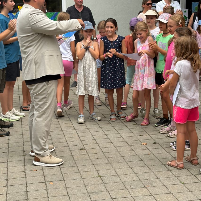 Eine Gruppe von Kindern und Erwachsenen steht in einem Kreis auf einem Ziegelboden, angeführt von einem Mann im Anzug. Die Kinder halten Papiere in der Hand und nehmen an einer Aktivität teil.