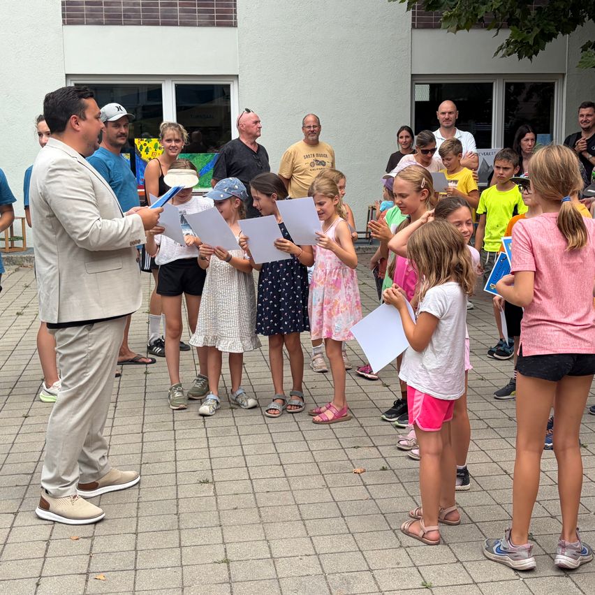 Ein Mann leitet eine Gruppe von Kindern bei einer Aufführung, hält Papiere in der Hand und spricht zu ihnen, während sie im Kreis stehen.