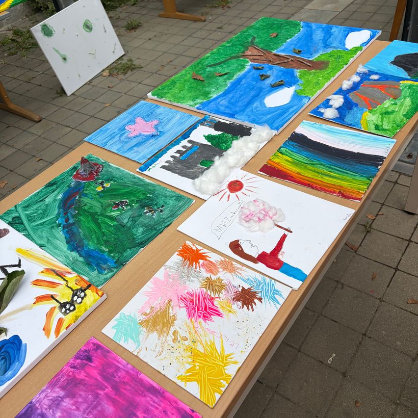 A wooden table displays a collection of colorful paintings, including landscapes, abstract designs, and a drawing of a person with a thought bubble.