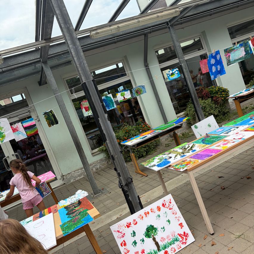 A group of children are painting on tables outside a building with glass walls. The tables are covered with colorful paintings.