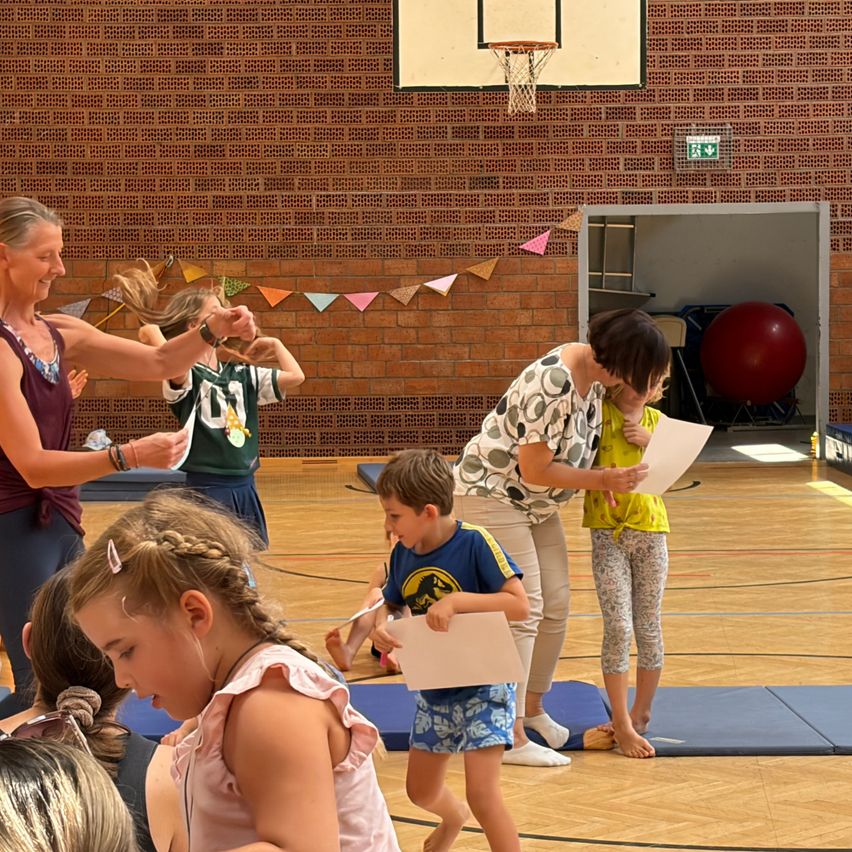 Eine Gruppe von Kindern und Erwachsenen versammelt sich in einer Turnhalle, einige halten Papiere in den Händen und andere sind in Aktivitäten verwickelt. Ein Basketballkorb ist an der Wand sichtbar.