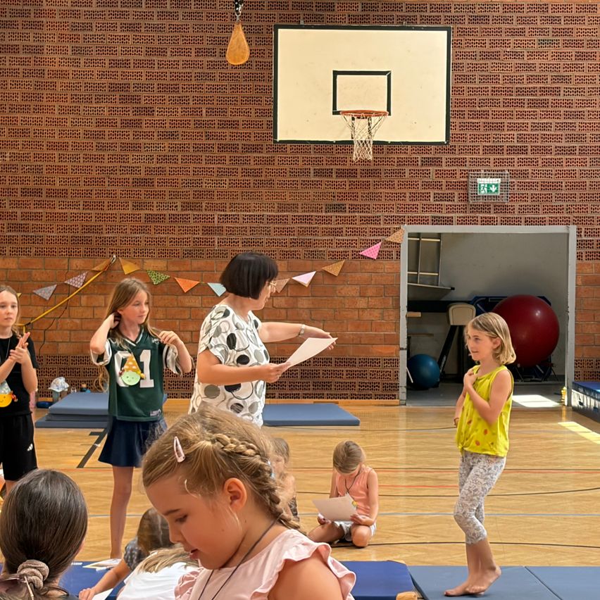 Eine Gruppe junger Mädchen und eine Frau stehen in einer Turnhalle, eine Mädchen liest von einem Papier. Die Halle hat eine Ziegelwand, einen Basketballkorb und bunte Dekorationen.