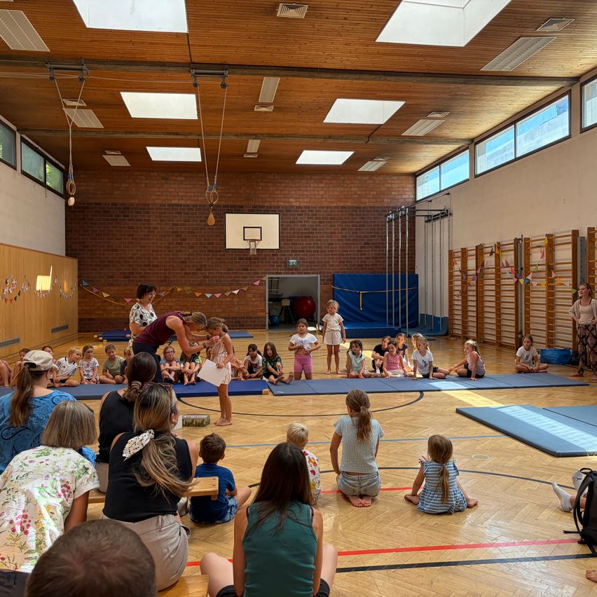 Eine Gruppe von Kindern und Erwachsenen versammelt sich in einer Turnhalle. Eine Frau steht in der Mitte und hält ein Stück Papier. Die Kinder sitzen auf blauen Matten auf dem Boden. Die Turnhalle hat Ziegelwände, einen Basketballkorb und verschiedene Trainingsgeräte.
