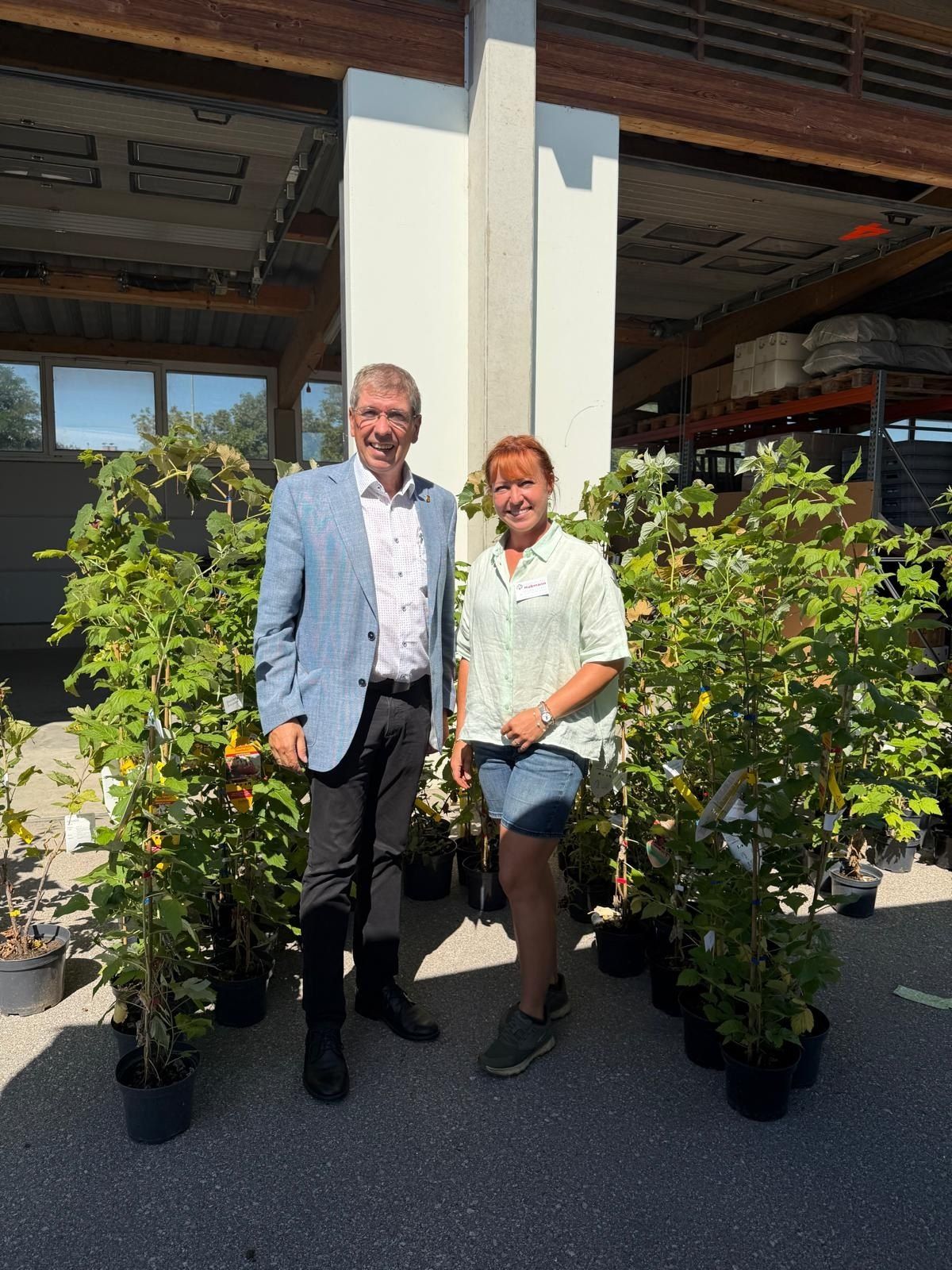A man and a woman stand next to potted plants in a greenhouse, smiling for the camera.