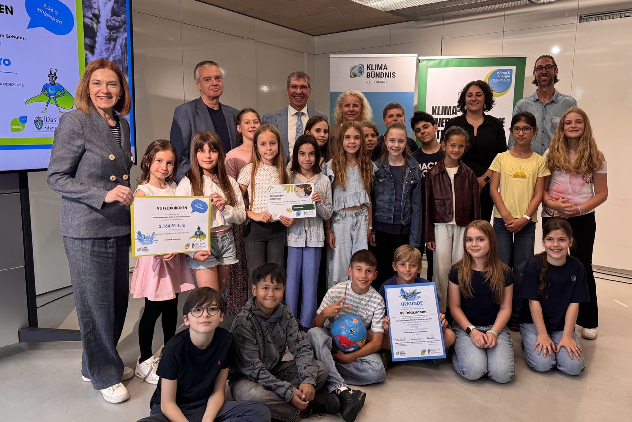 Gruppe von Kindern und Erwachsenen steht für ein Foto, einige halten Urkunden. Ein Banner hinter ihnen lautet 'Klima Bundnis'.