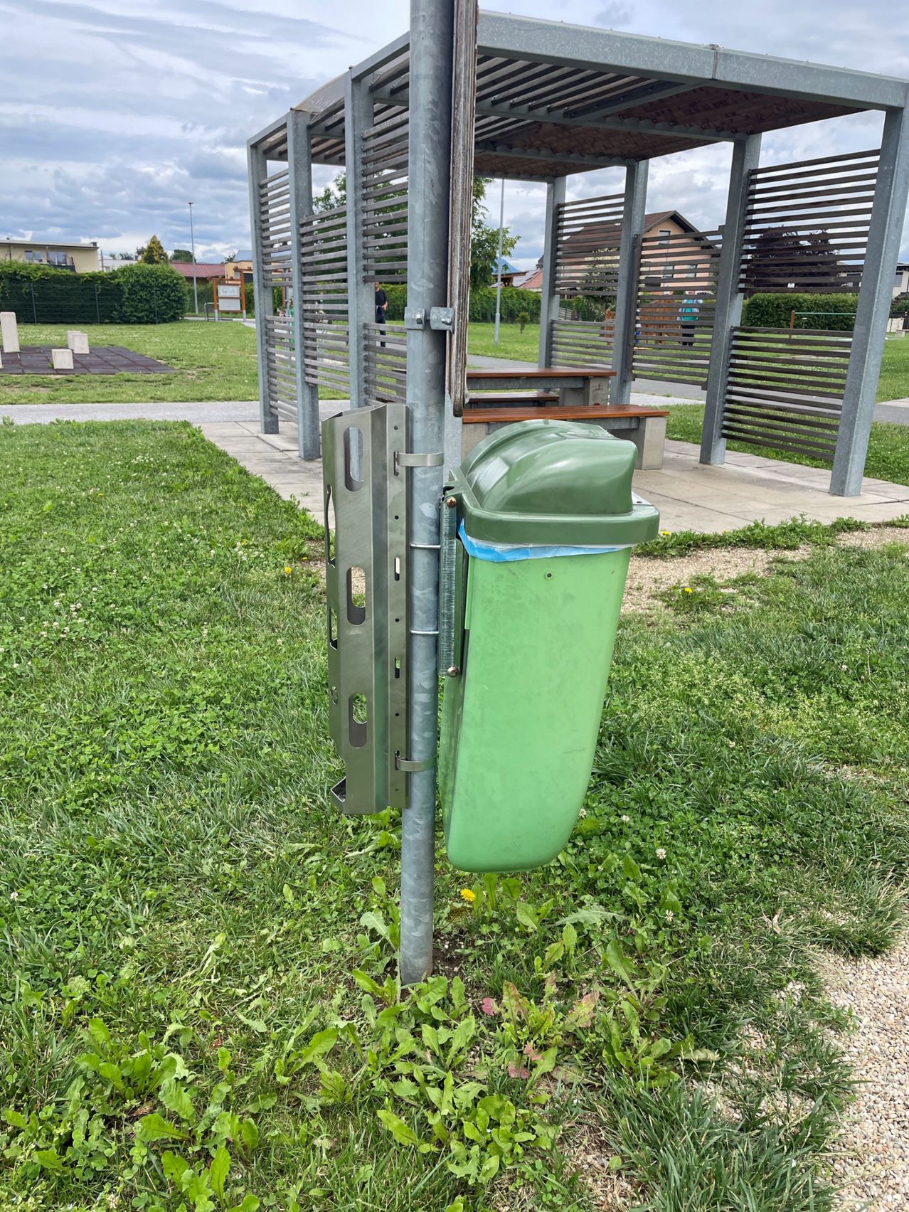Ein grüner Mülleimer ist an einem Metallpfosten in einem Grasbereich befestigt, mit einem Schuppen und einer Bank im Hintergrund.