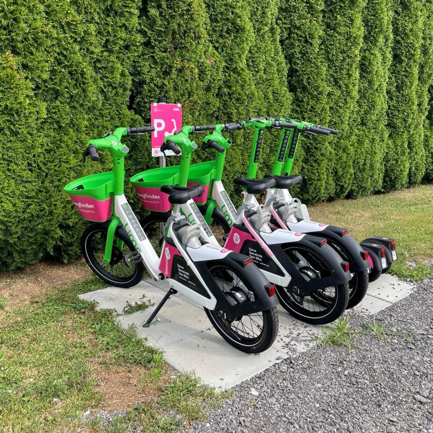 Vier grüne und rosafarbene Elektrofahrräder sind in einer Reihe geparkt, hinter ihnen befindet sich ein rosa Schild.