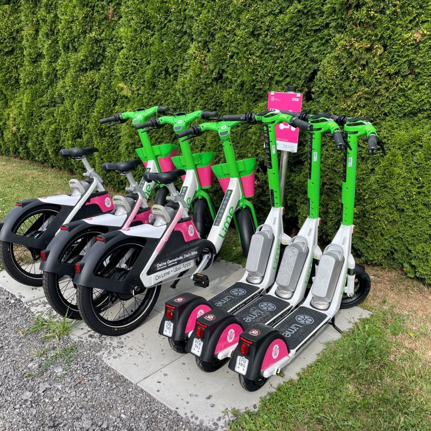 Eine Reihe von Elektrorollern, die auf einem gepflasterten Bereich mit einem Hecke im Hintergrund aufgereiht sind. Die Roller sind grün und rosa mit weißen Sitzen.