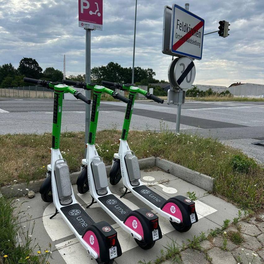 Drei Lime Elektroroller stehen auf einer Betonfläche in der Nähe einer Straße mit Ampeln und einem Schild.