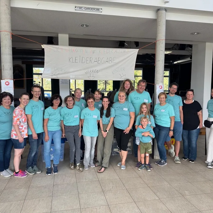 Eine Gruppe von Menschen in hellblauen Shirts posiert für ein Foto in einem Raum mit einem Banner, das 'Kleiderabgabe' besagt.