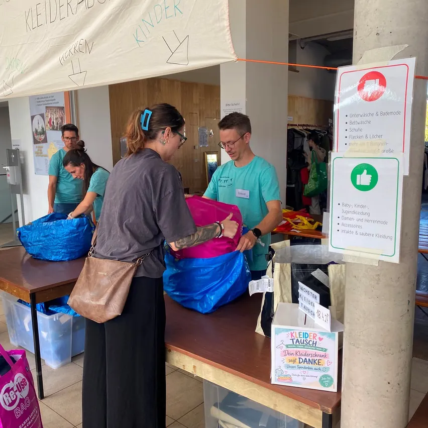 Eine Gruppe von Menschen sortiert durch Taschen mit Kleidung in einem Raum mit einem Banner, das 'Kleiderabgabe Kinder' sagt. Eine Frau hält eine blaue Tasche und ein Mann hält eine rosa Tasche. Es gibt Lagerboxen und ein Schild mit Anweisungen auf der rechten Seite.