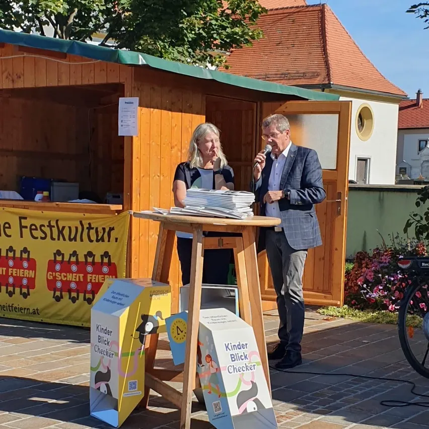 Ein Mann und eine Frau stehen vor einer hölzernen Hütte. Der Mann hält ein Mikrofon und scheint zu sprechen, während die Frau neben ihm mit einem Stapel Papiere steht.
