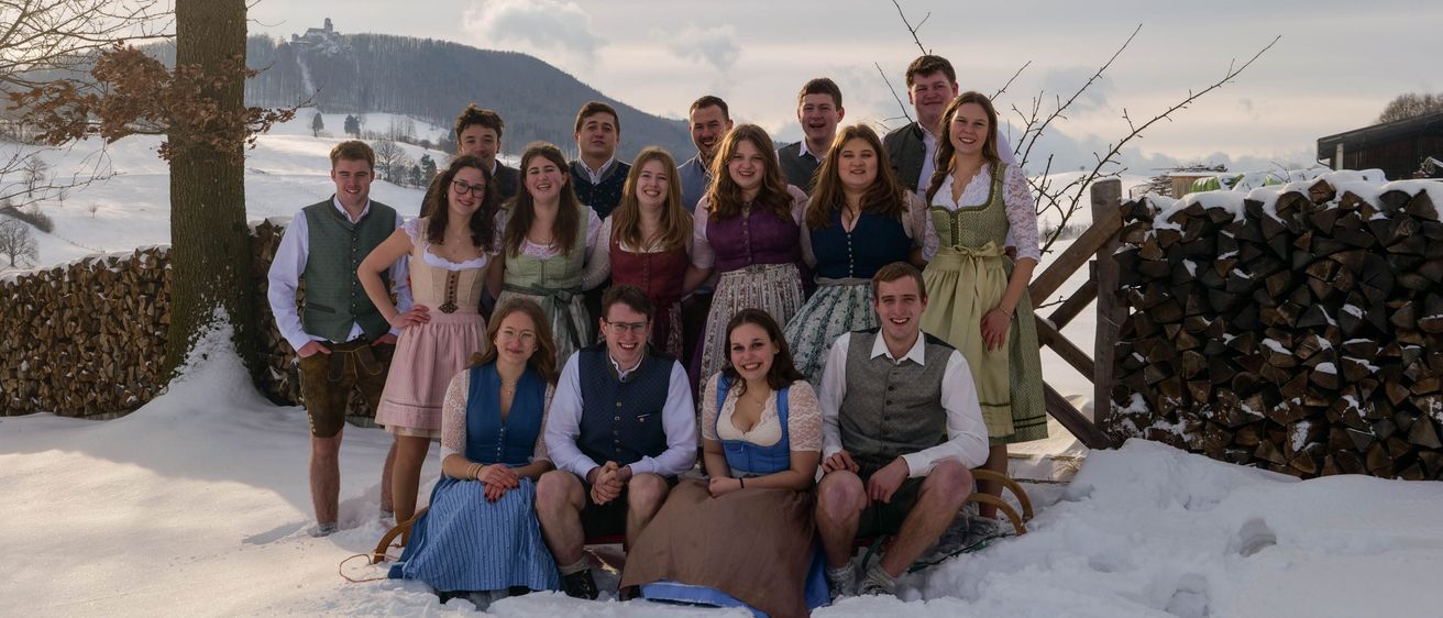 Eine Gruppe von Menschen in traditioneller bayerischer Kleidung posiert für ein Foto im Schnee mit einem Berg im Hintergrund.