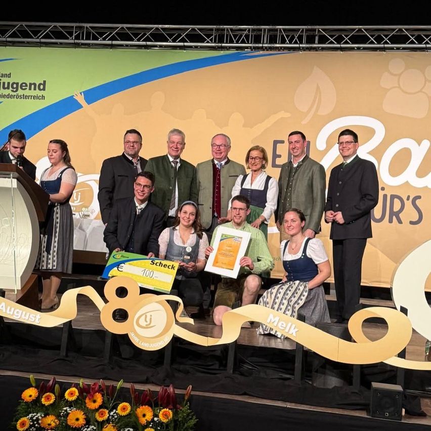 Eine Gruppe von Menschen steht auf einer Bühne, hält Urkunden in den Händen, lächelt und posiert für ein Foto. Der Hintergrund hat ein Banner mit Text und Logo.