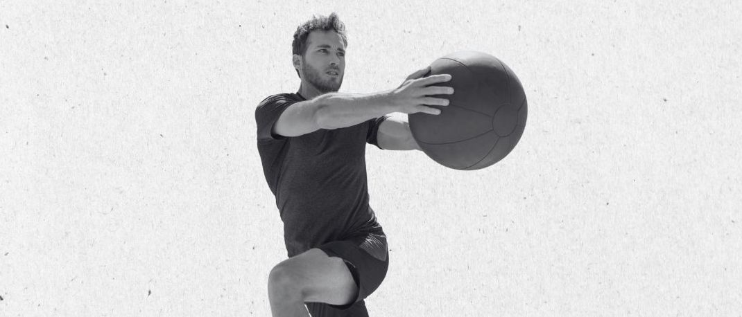 Ein Mann in Sportbekleidung springt und hält einen Basketball in beiden Händen, mitten in der Luft mit konzentriertem Ausdruck.
