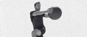 Ein Mann in Sportbekleidung springt und hält einen Basketball in beiden Händen, mitten in der Luft mit konzentriertem Ausdruck.