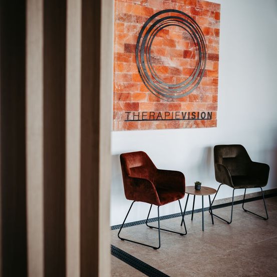 Ein roter Stuhl und ein kleiner Tisch mit einer Pflanze stehen neben einer Wand mit Ziegeldesign und THERAPIEVISION-Schild.