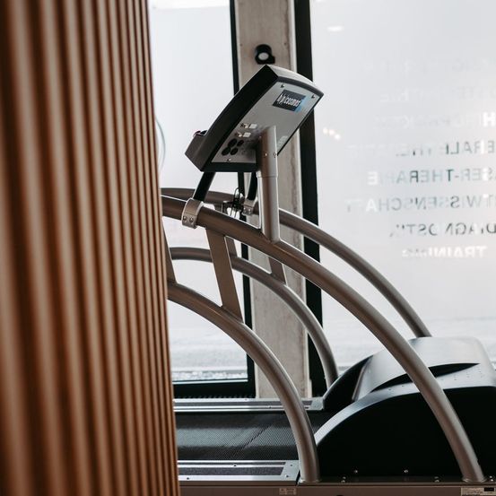 Ein Laufband steht in einem Fitnessstudio mit Glastür und schwarzer Matte. Das Laufband hat ein Bedienfeld und ein digitales Display.