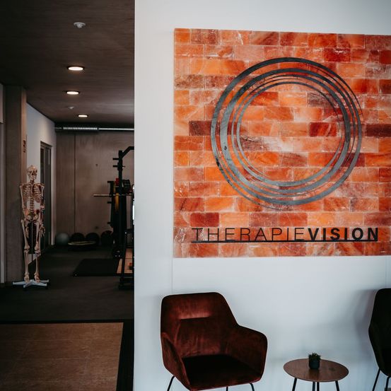 Ein Innenraum mit einer großen Ziegelwand, auf der 'THERAPIEVISION' steht. Ein Skelettmodell ist links positioniert, während Fitnessgeräte und Bälle im Hintergrund zu sehen sind. Ein brauner Stuhl und ein kleiner Tisch mit einer Pflanze stehen im Vordergrund.