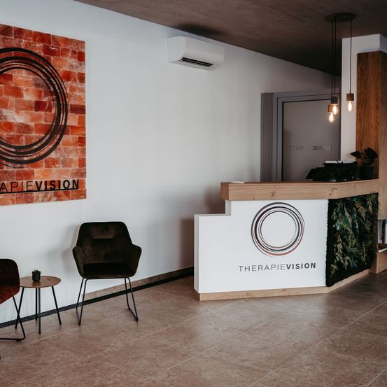 Ein Raum mit einer weißen Wand, einem schwarzen Stuhl und einem Tisch mit einer Pflanze. Eine hölzernen Rezeptionstresen mit einer grünen Wand auf der rechten Seite. Eine Klimaanlage ist an der Wand montiert. Das Wort 'THERAPIEVISION' steht auf dem Tresen.