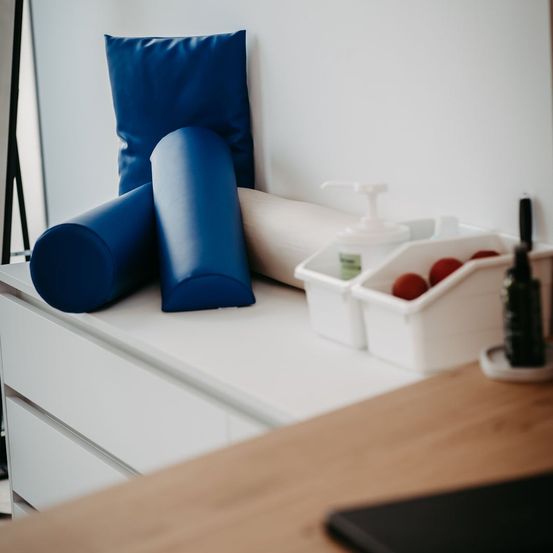 Ein weißer Anzugschrank beherbergt mehrere blaue Kissen, eine weiße Schaumstoffrolle und eine Lotionsflasche. Daneben enthält ein weißer Behälter rote Bälle. Ein schwarzer Pfefferstreuer und ein Tablet befinden sich auf der Arbeitsplatte.