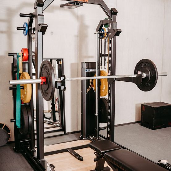Ein Fitnessstudio-Setup mit einer Langhantel und Gewichten, einer Bank und Spiegeln. Verschiedene farbige Gewichte hängen an Ständern.