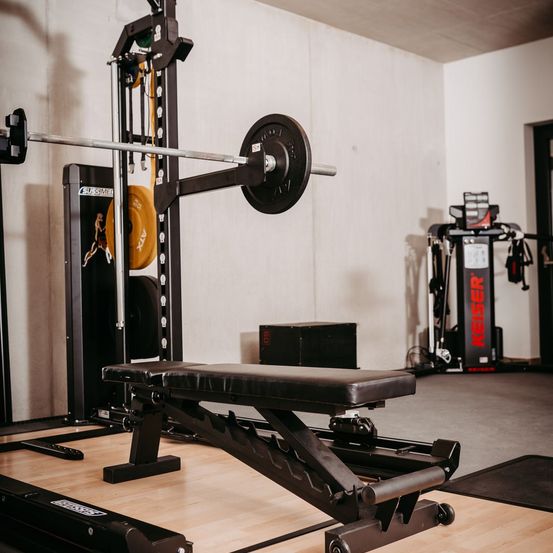 Ein Fitnessstudio mit einer Langhantel, Gewichten und einer Bank. Das Fitnessstudio ist mit einem Gewichtsregal, einer Matte und einer Wand mit einem Keiser-Logo ausgestattet.