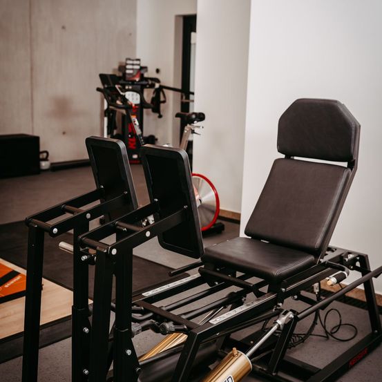 Ein Fitnessstudio-Interieur mit einem schwarzen Trainingsgerät mit zwei Sitzen und gepolsterter Rückenlehne. Im Hintergrund befinden sich ein rotes Fahrrad und verschiedene Fitnessgeräte.