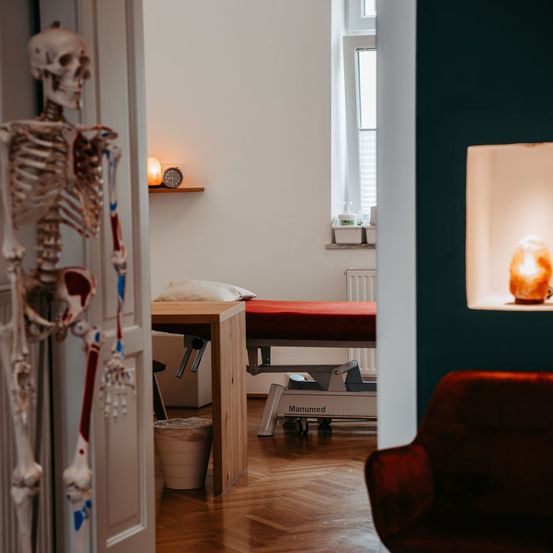 Ein Innenansicht eines Raumes mit einem menschlichen Skelettmodell, einem Massagetisch, einem roten Stuhl und einer beleuchteten Salzlampe. Der Raum hat Holzböden und weiße Wände.