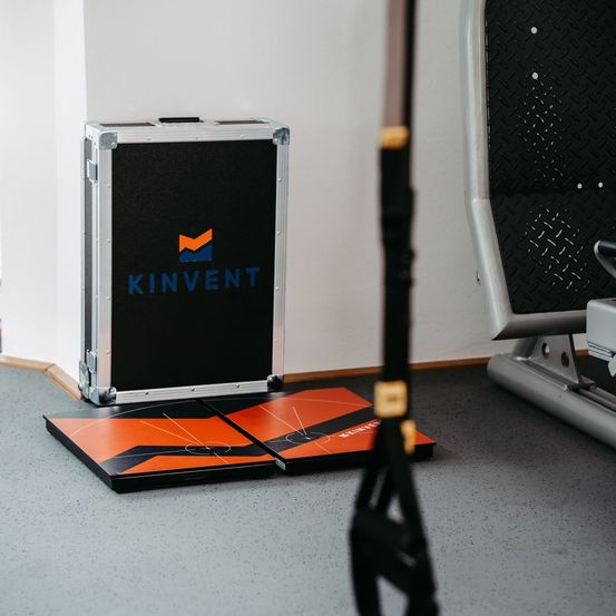 Ein Kinvent-Koffer mit orange-schwarzem Design steht auf dem Boden in der Nähe von Fitnessgeräten.