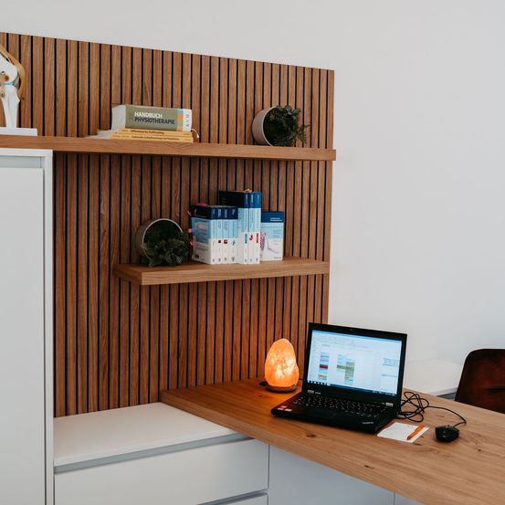 Ein Holzschreibtisch mit Laptop, Maus und Notizblock. Eine Lampe steht auf dem Schreibtisch. Ein Schrank mit Regalen ist an der Wand befestigt. Bücher und Topfpflanzen stehen auf den Regalen.