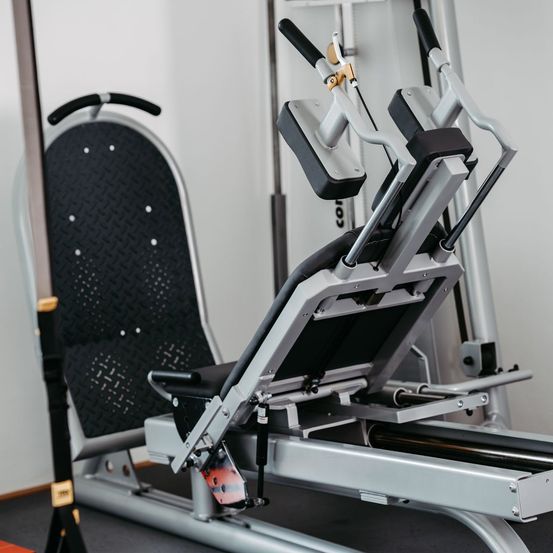 Eine Nahaufnahme eines modernen Krafttrainingsgeräts mit schwarzen und grauen Teilen. Es hat einen großen Sitz und Griffe für Arm- und Beinübungen. Das Gerät steht auf einem Holzboden, mit einer weißen Wand im Hintergrund.