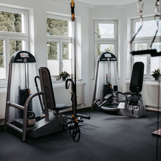 Ein Fitnessstudio mit weißen Wänden, drei Trainingsgeräten und großen Fenstern, die natürliches Licht hereinlassen. Ein Heizkörper ist unter den Fenstern sichtbar.
