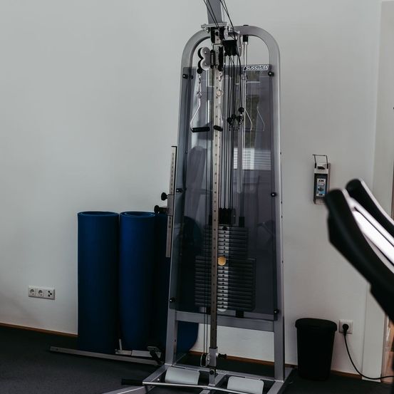 Ein Fitnessstudio-Innenraum mit einer Gewichtsmaschine, zwei blauen Zylindern, einem Mülleimer und einer Steckdose. Das Fitnessstudio hat weiße Wände und einen schwarzen Boden.