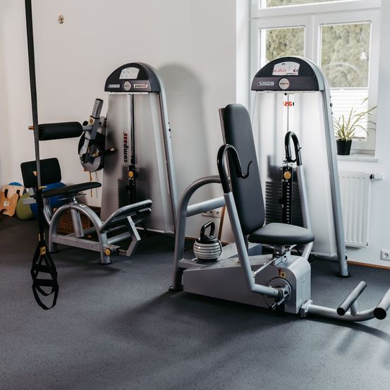 Ein Indoor-Fitnessstudio verfügt über verschiedene Trainingsgeräte. Eine Hantelglocke steht vor einem Rudergerät. Ein Topf steht auf der Fensterbank.