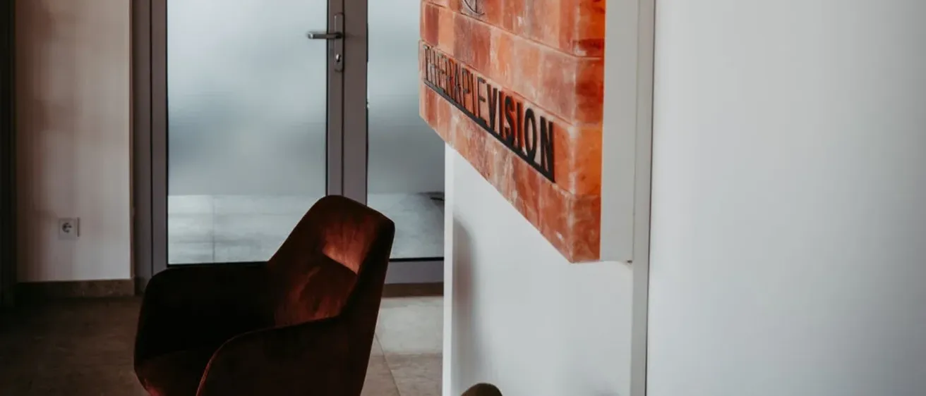Ein Büroraum mit einer Glastür, einer Ziegelwand mit einem 'Fernsehen'-Schild und zwei Stühlen. Der Boden ist gefliest und die Wand weiß.